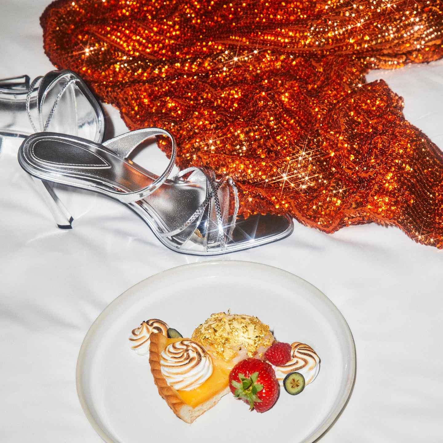Dessert on plate, silver heels, and orange glittery blouse laying on a white bed at Sanderson hotel London