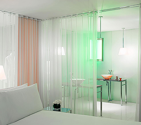 Bathroom with green light behind white curtain with corner of white bed in foreground