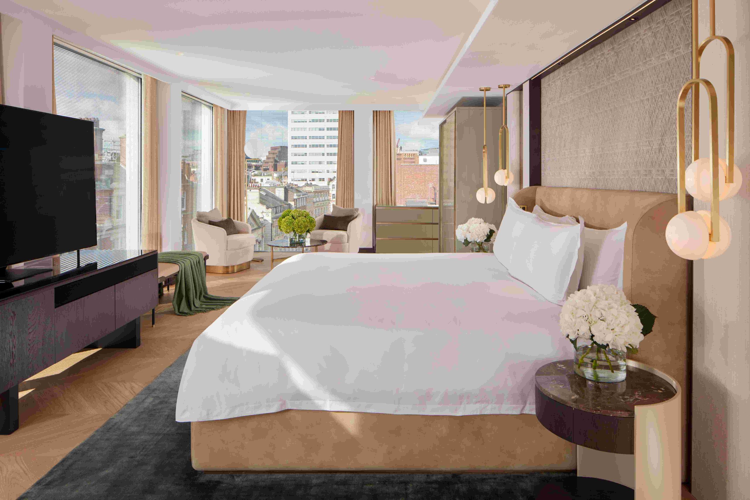King bed with white linens, dark wooden bedside table, pendant lights, white chair and close in the background with large windows overlooking London buildings