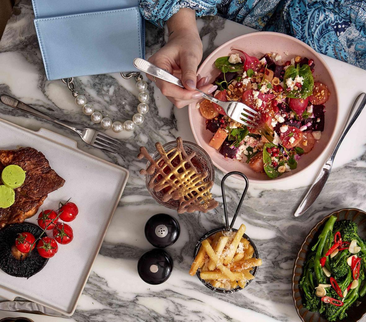 Salad in a pink bowl, blue purse, steak, fries, greens, martinis on a marble table