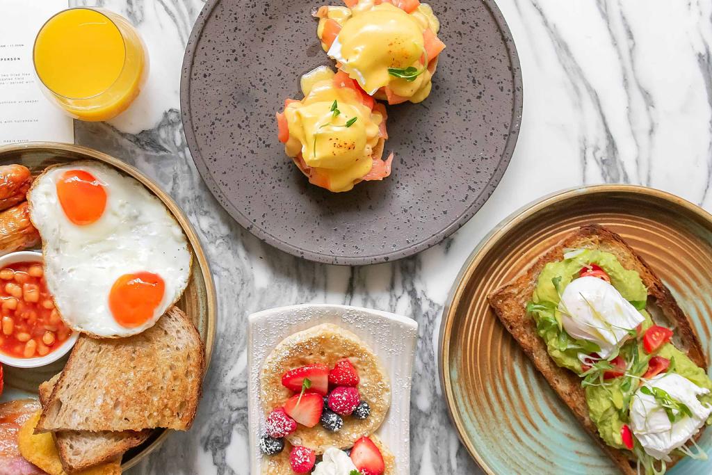 Marble table with delicious breakfast dishes, English breakfast, pancakes, poached eggs on English muffin and mashed avocado on sourdough bread.