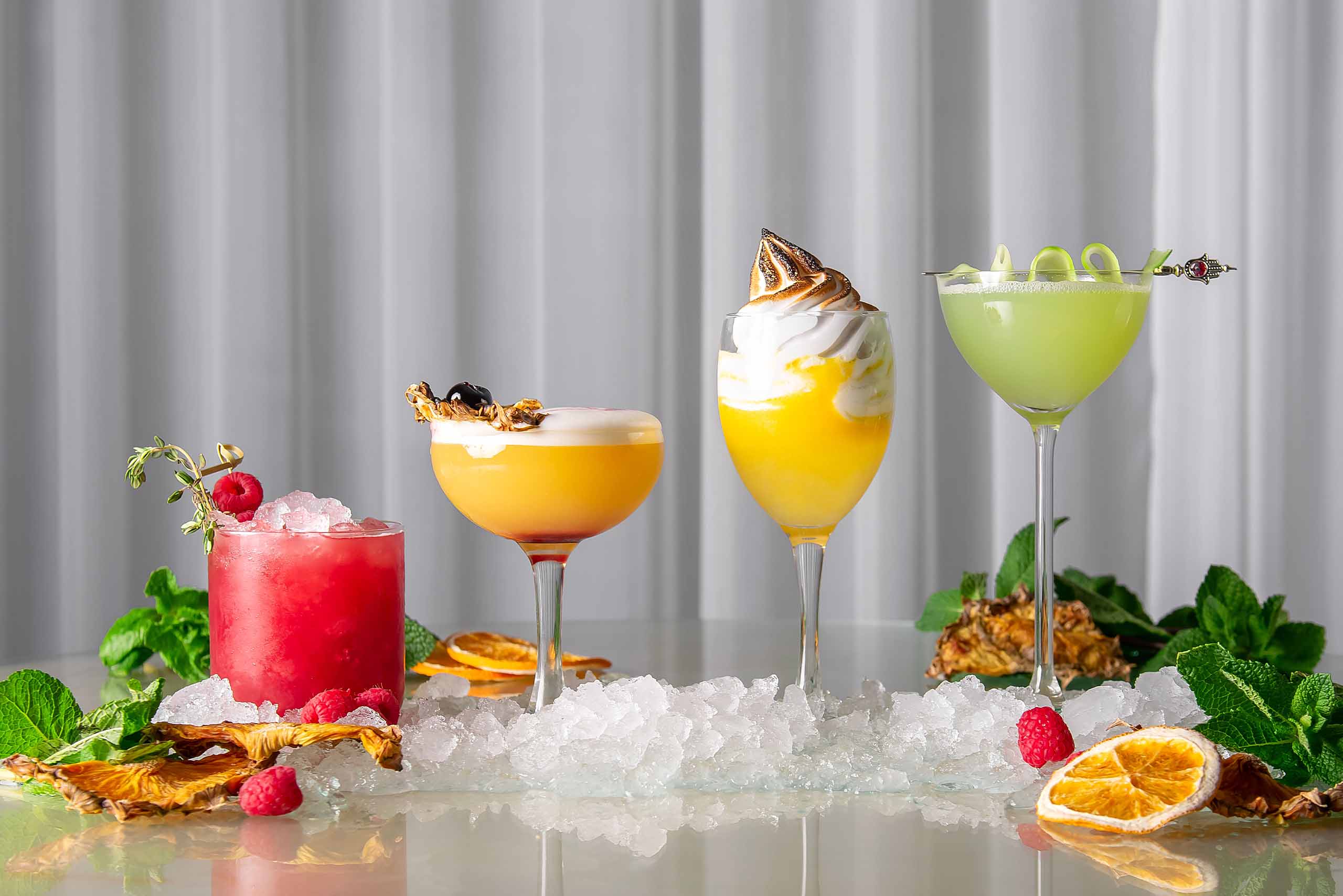 A row of colourful cocktails with fruity garnish on a table decorated with fruits and herbs.