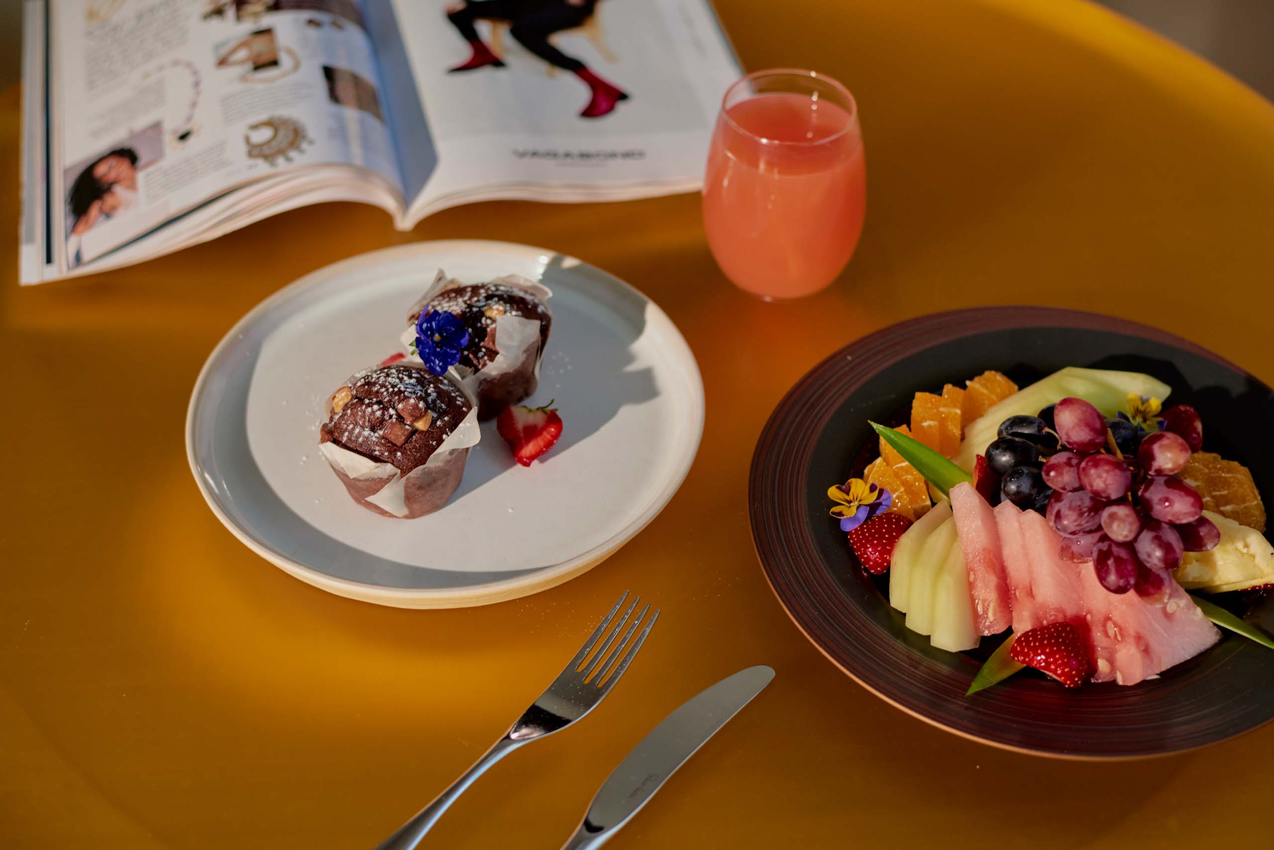 A table displaying a colourful arrangement: a dish of freshly cut fruits, a plate of homemade muffins, a glass of pink beverage, and an open book invitingly set for a cosy and relaxed moment.