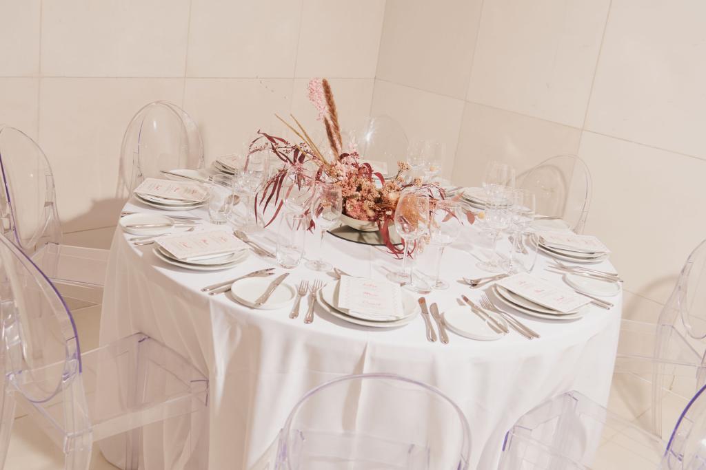 The table is decorated with a white tablecloth and wedding centre piece s surrounded by clear chairs. 