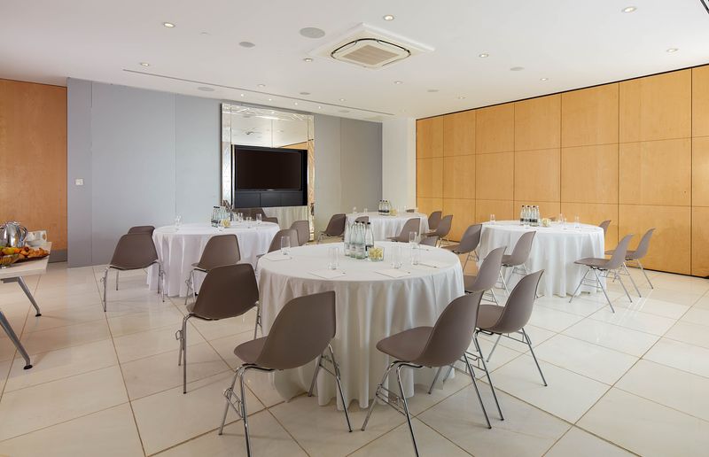 Boardroom with round tables with white tablecloths and gray chairs with a tv screen at the front of the room