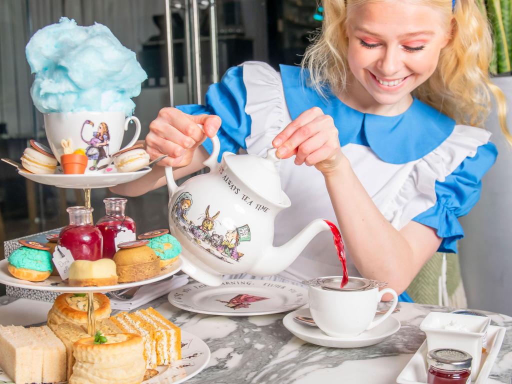 Woman in Alice in Wonderland costume pours tea into cup and a tea tray with food is in the foreground