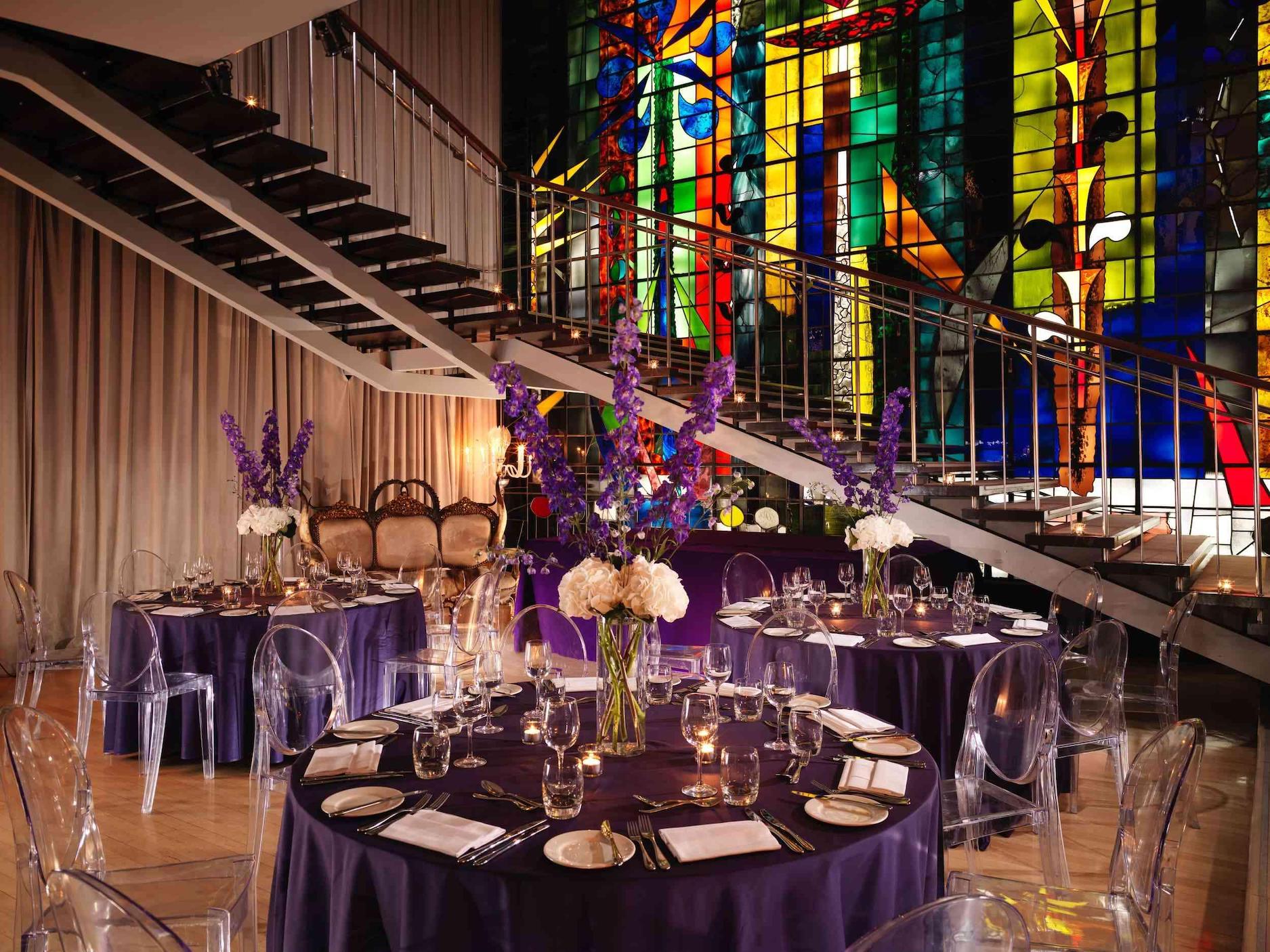 Large room with glass stained windows and round tables with purple tablecloths and large bouquet centerpieces