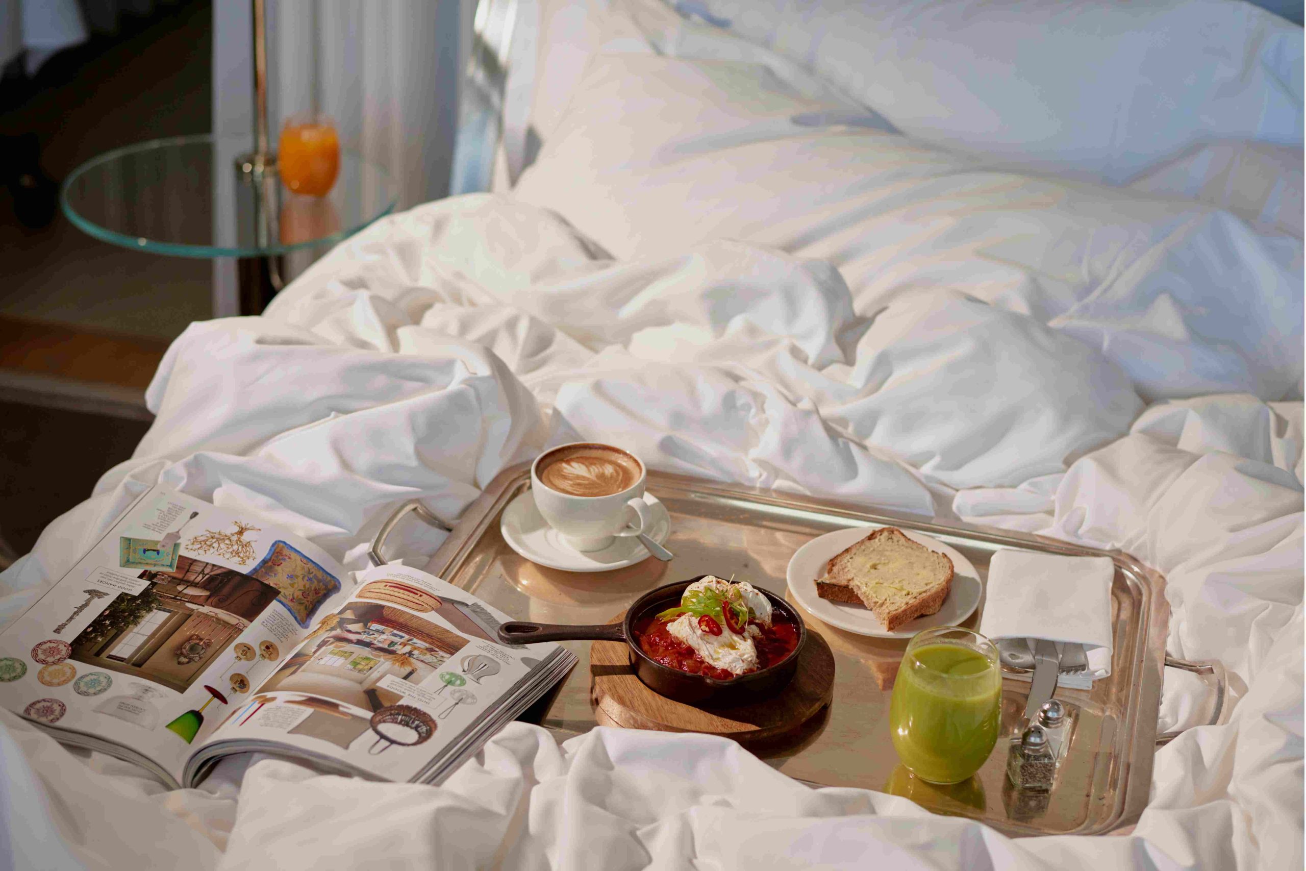 Coffee, toast, eggs, green juice on a tray with a magazine open on a white bed