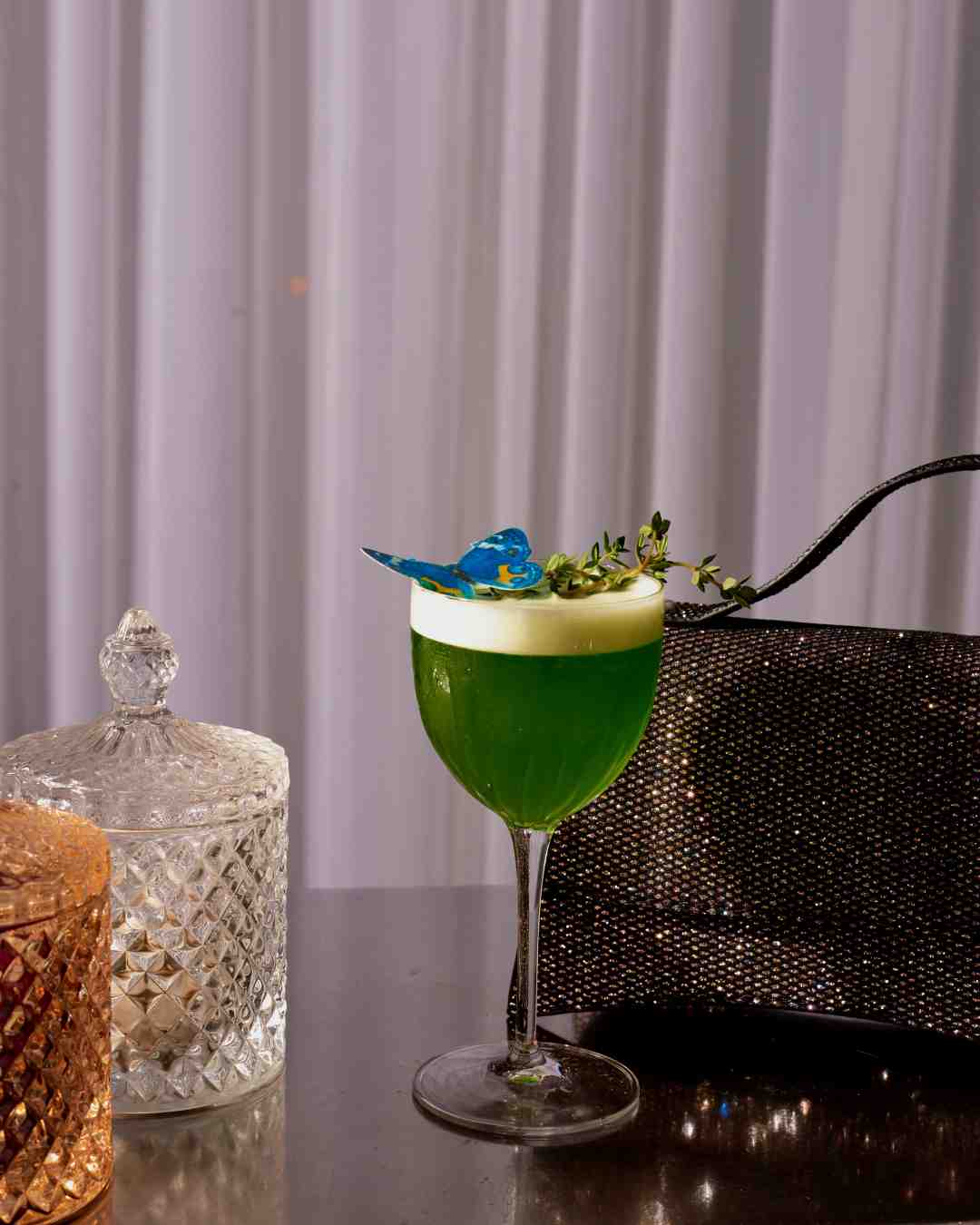 Green cocktail and small grey handbag with glassware on a silver bar table with white curtains in the background