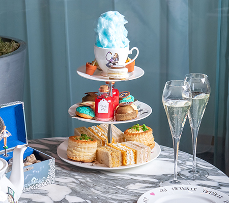 Elaborate tea tray with pastries, sandwiches, scones, and a tea cup on top with cotton candy, champagne glasses filled with champagne to the side of the tea tray