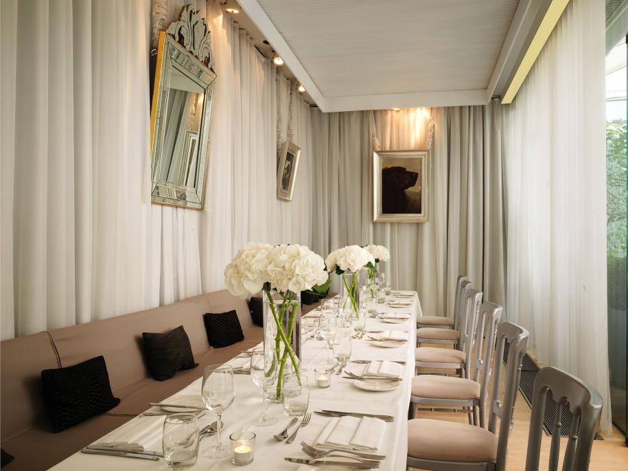 Room with beige curtains and long rectangular table with white tablecloth, beige booth with black pillows, glassware, utensils, and large white flowers on table