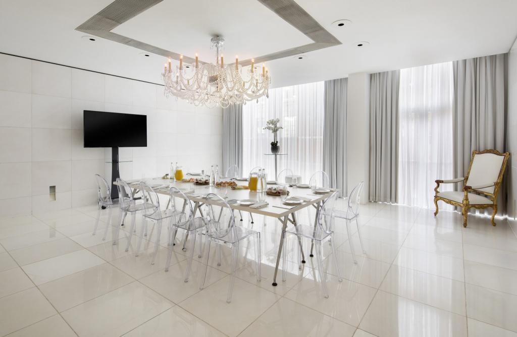 Large bright room with white floors, ceilings, and curtains with elaborate chandelier and large rectangular table and clear chairs surrounding