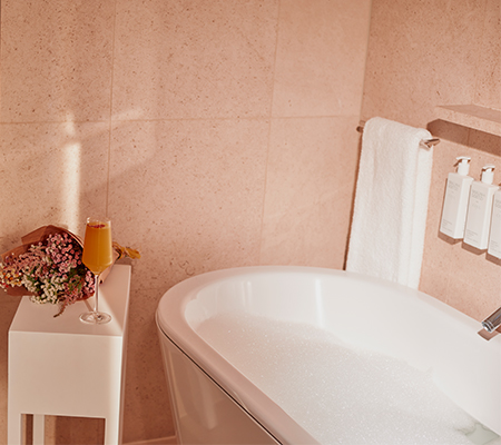 White bathtub filled, white towel hanging, white side table with bouquet of flowers and glass of mimosa
