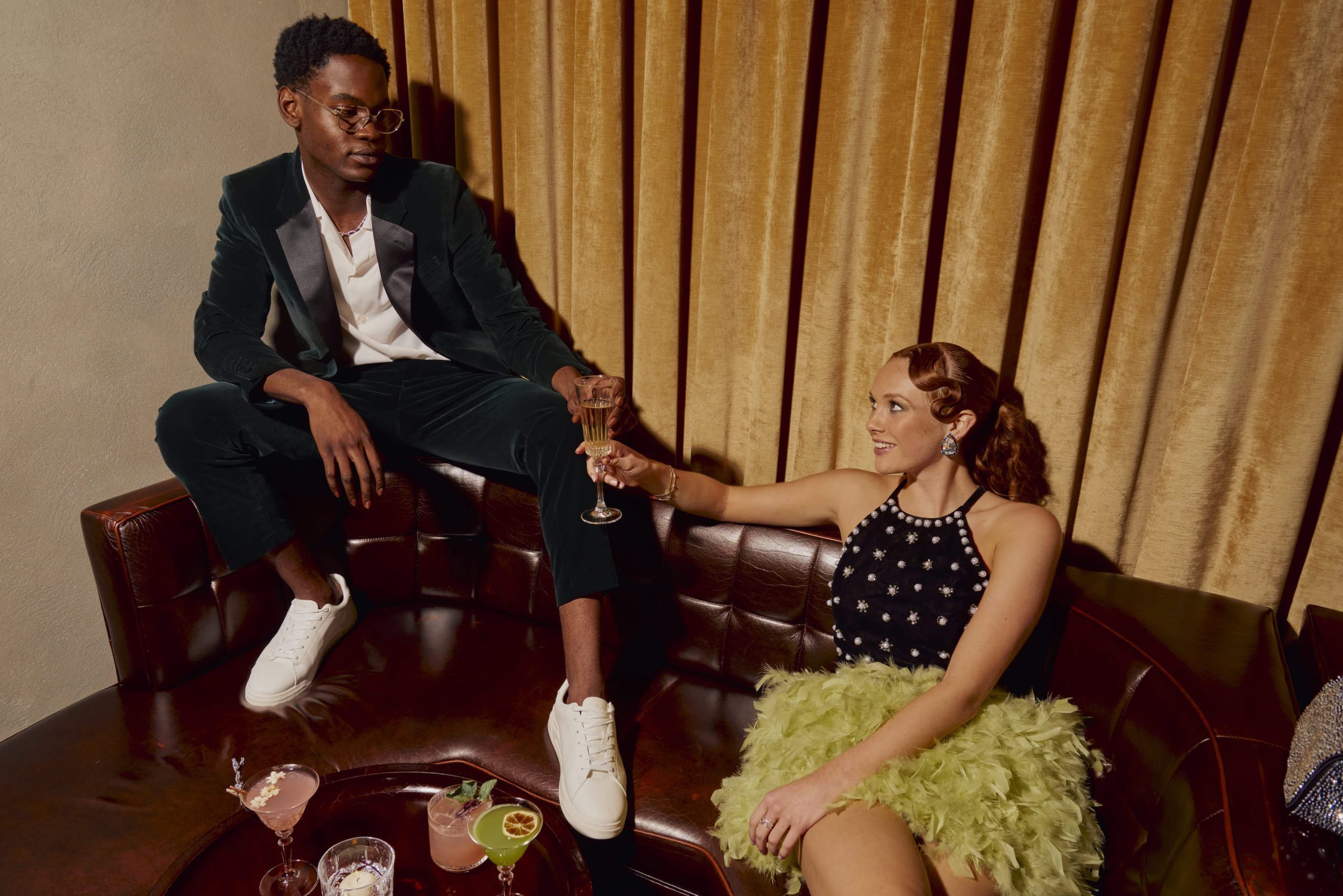 Man and woman on leather sofa holding a champagne glass looking at each other with beige curtain in the back