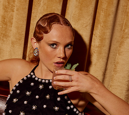 Red-headed woman with large earrings and black top with jewels puts cocktail up to her mouth