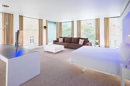 Large room with white bed, brown sofa in corner, white ottoman and large floor to ceiling windows