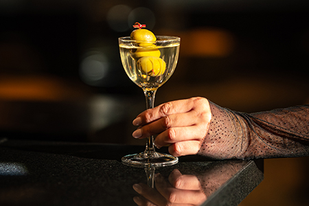 Martini with olives sitting on a bar with hands holding it