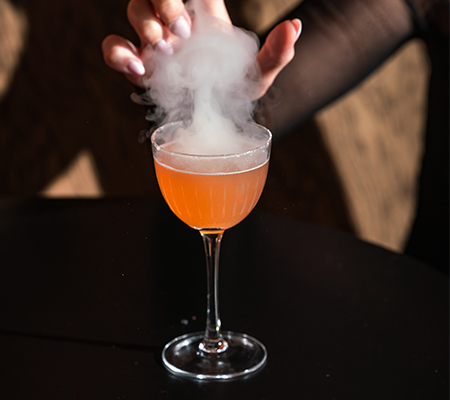 Orange cocktail with smoke rising above it and hand over top it on black countertop
