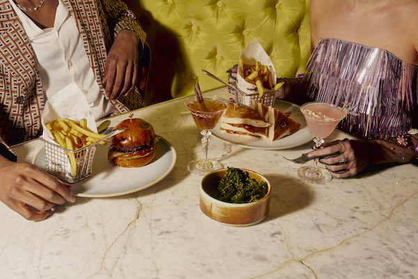 Woman in glittery top holds onto pink cocktail and man holds onto plate with burger and fries