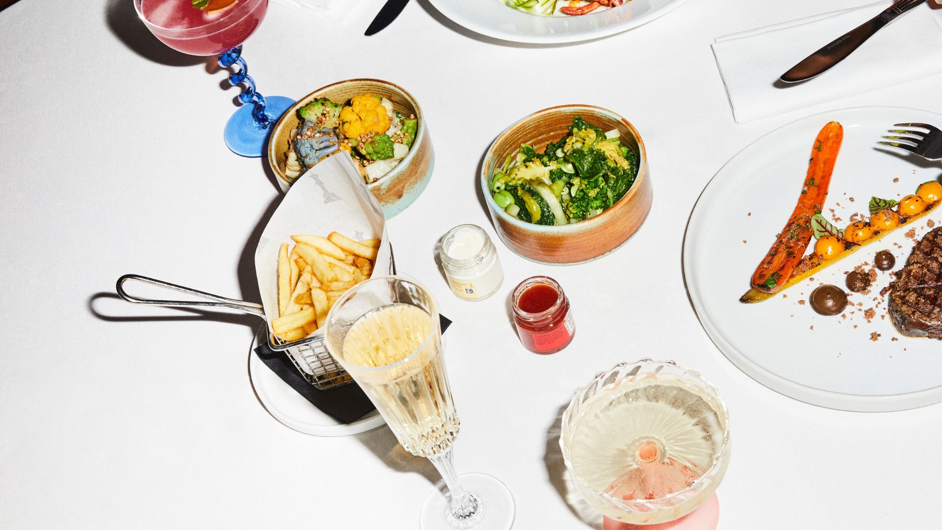 Fries, champagne, greens, steak, and other cocktails on a white table