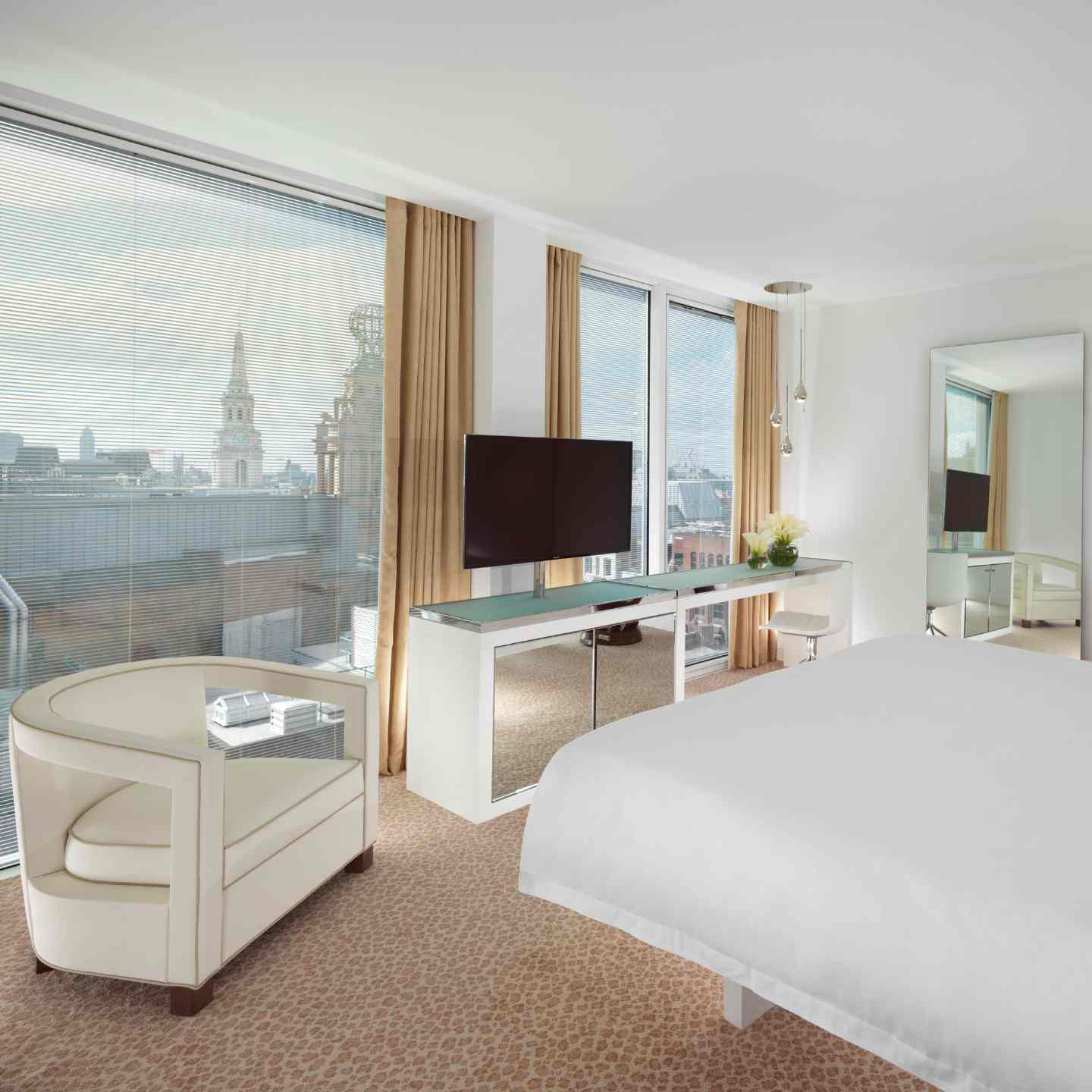 Hotel room with large white bed, white leather chair, white entertainment center with black TV, and floor-to-ceiling windows with views of London