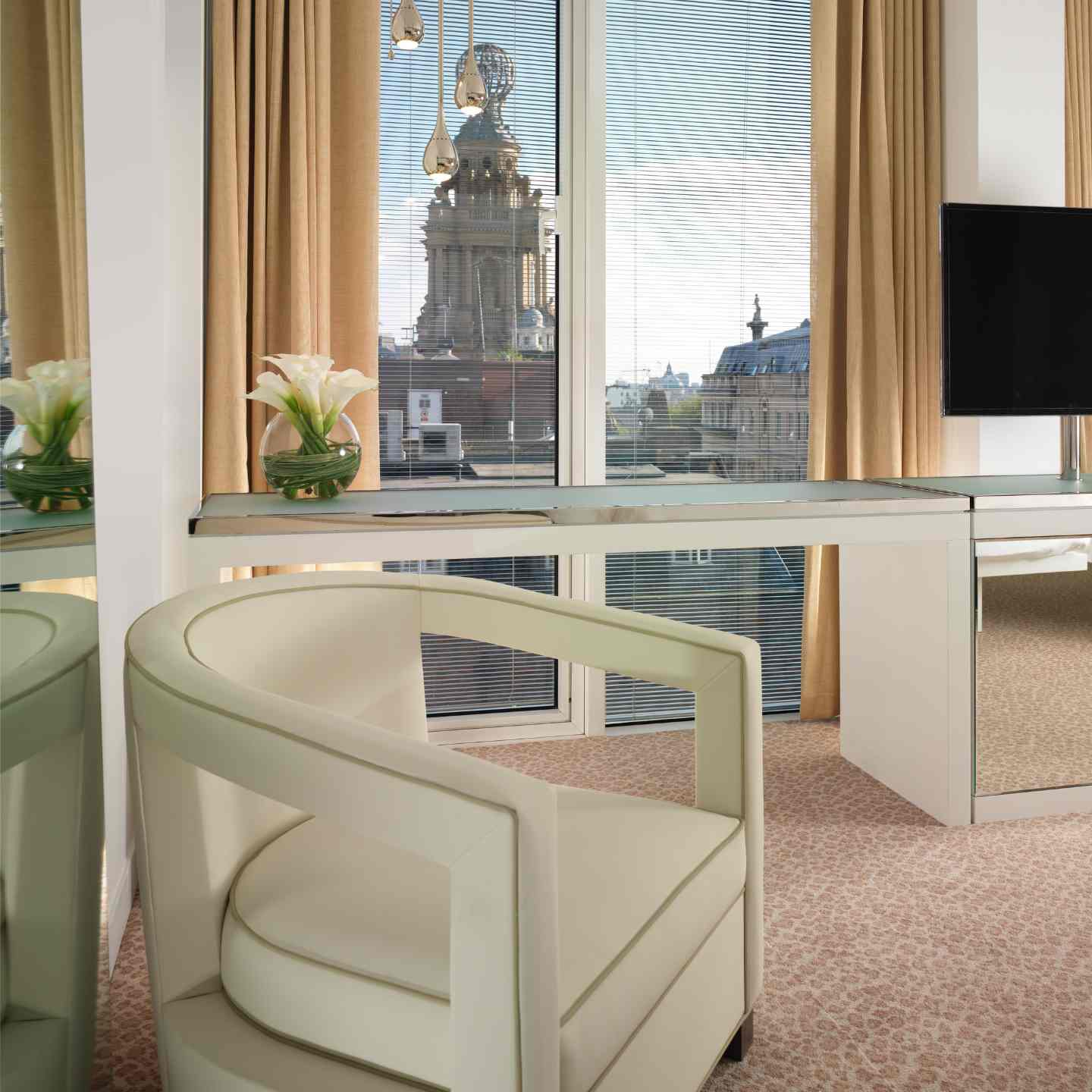 White leather chair with studs and white desk with flowers on it, floor-to-ceiling windows with beige curtains and views of London