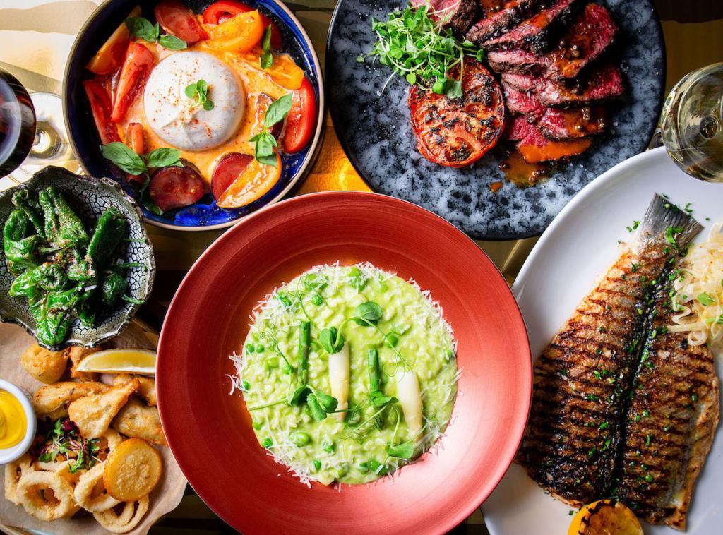 Green risotto in a red dish, grilled fish on a white plate, steak and tomatoes on a blue plate, and other dishes in the background on a table