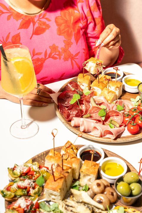 Focaccia, cured meats, olives, and tomatoes on wooden trays with a glass of sangria in the foreground, a woman in a pink and red flowered blouse int he background