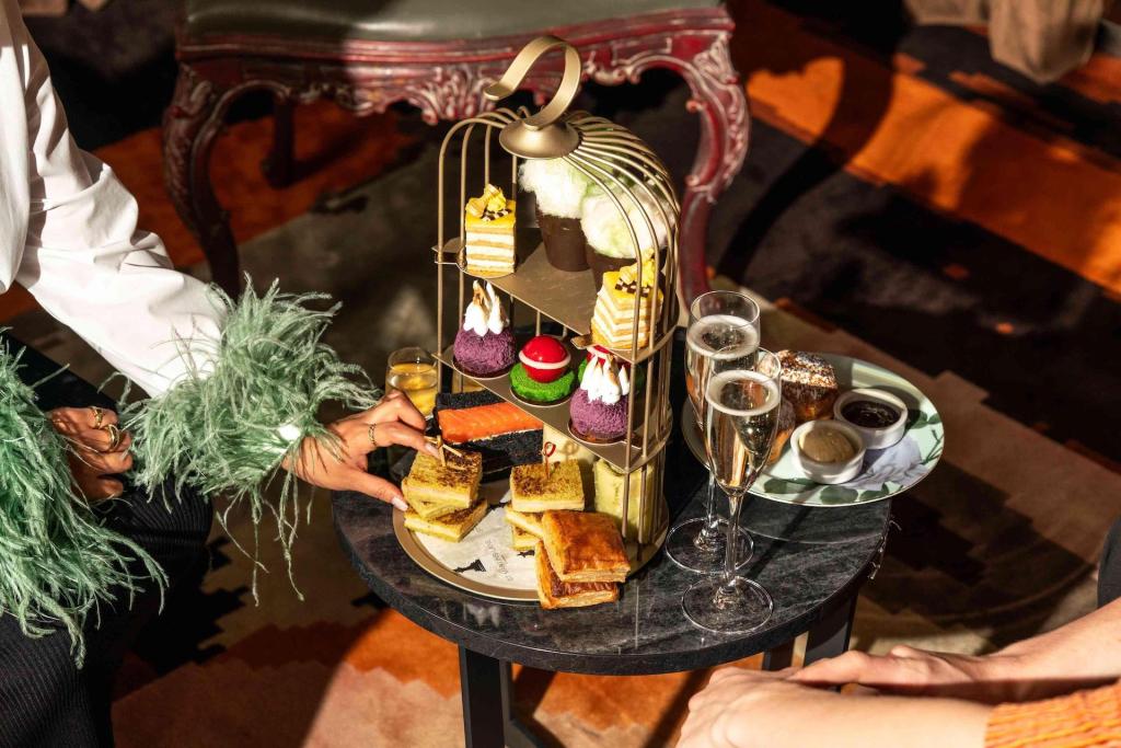 Brass tea tray with pastries, champagne flutes, and woman's hand in green blouse with feathers grabbing a dessert off the table