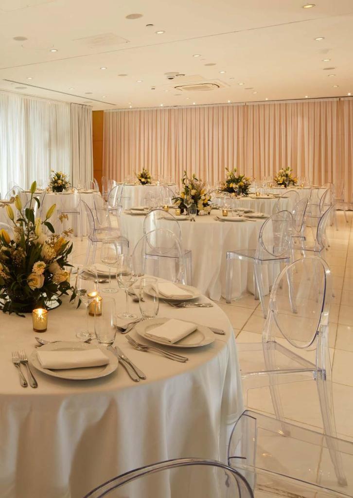 Banquet room with large white cloth tables and flowers as centerpieces with clear chairs surrounding the tables