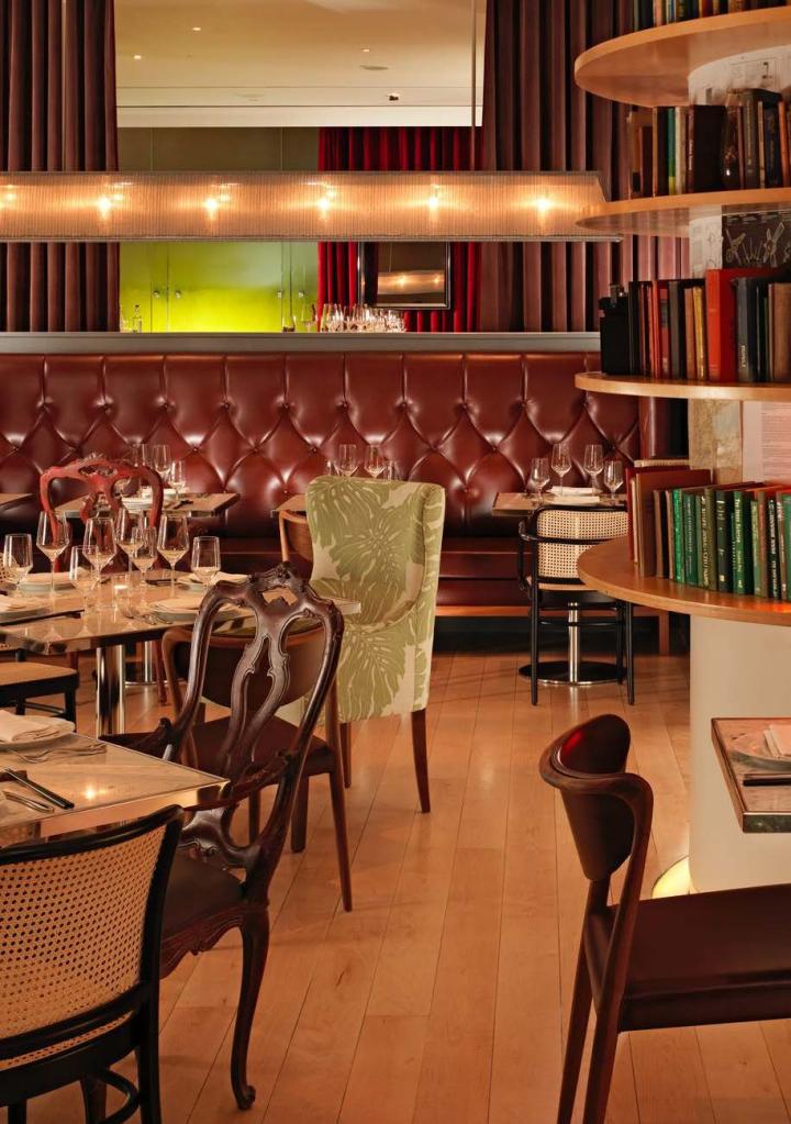 Restaurant room with tables and chairs of different sizes and tufted round banquette seating and bookcase in the background