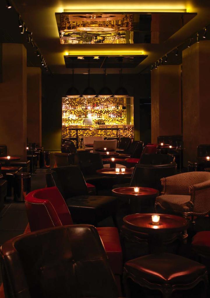 Dark lounge room with lounge chairs, small wooden tables with candles on it, and yellow lit bar in the background