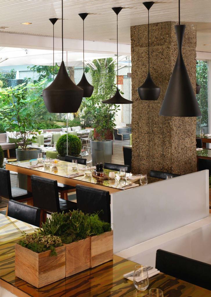 Restaurant space with black pendant lights, light brown rectangular wood tables, black chairs, and green plants throughout