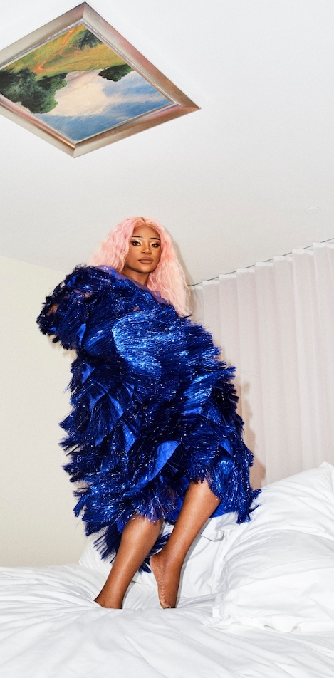 Woman with pink hair and wearing ruffly blue dress stands on top of white bed