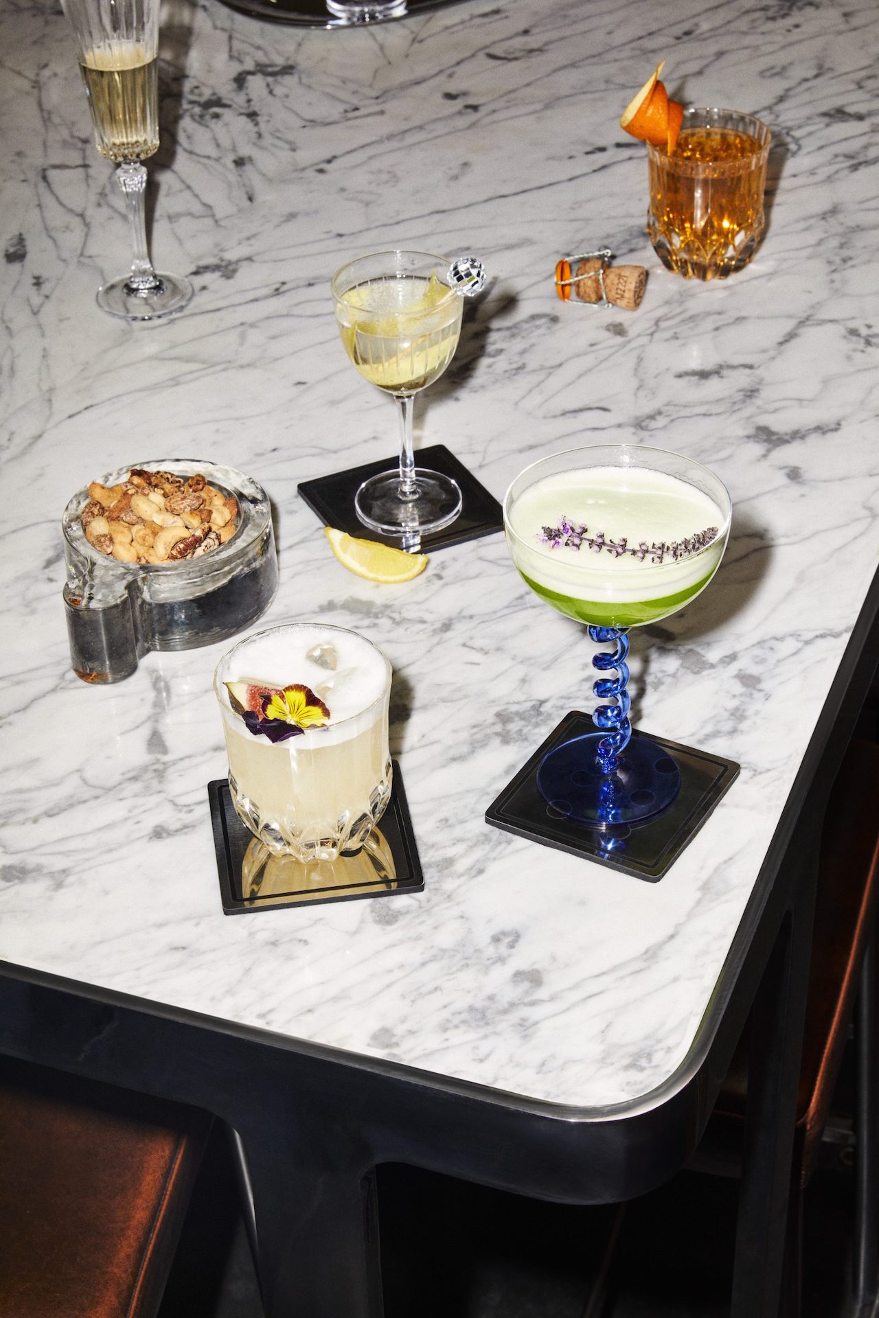 Five cocktails in various glasses on a marble tabletop with a bowl of nuts