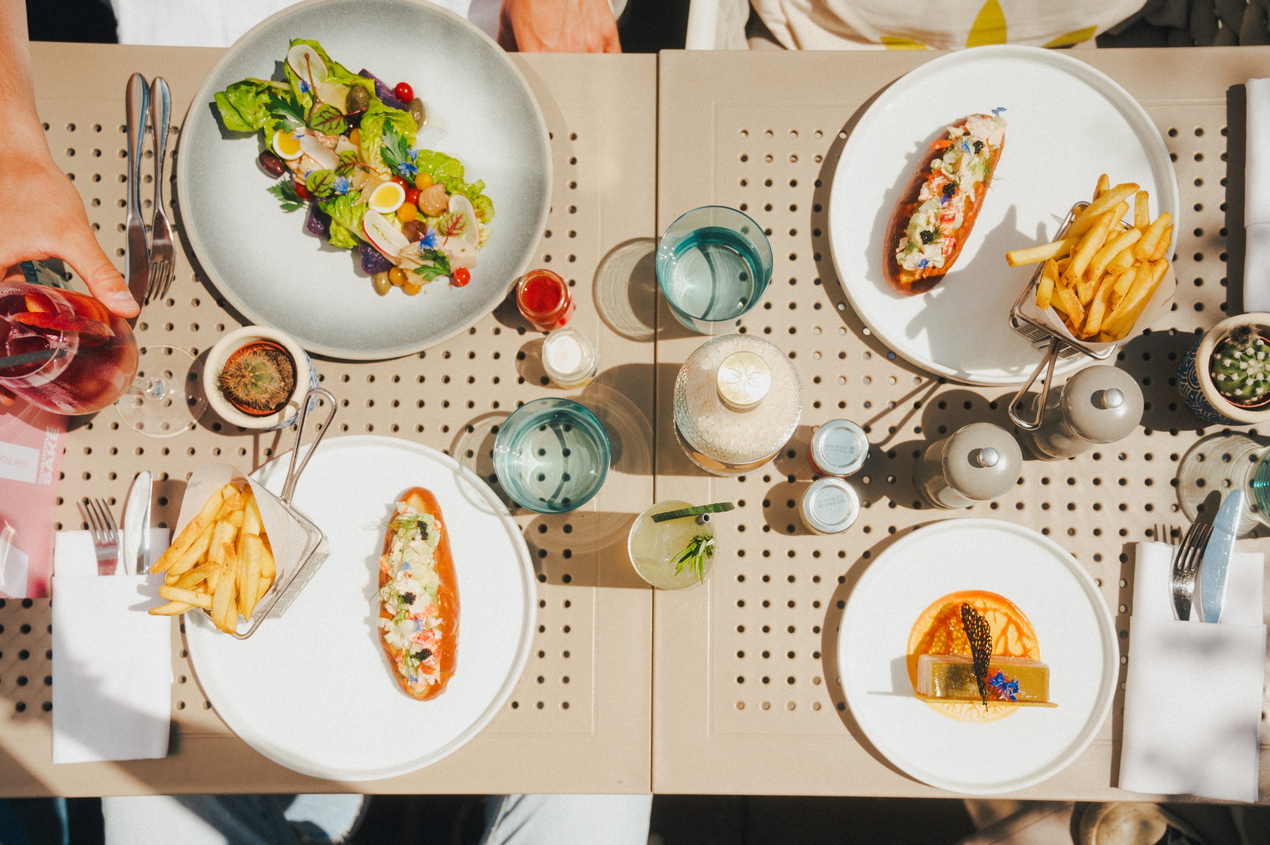 Above image of a table with four plates, two water glasses, four sauces, salt and pepper and a bottle. The plates have colorful food on them, one with a green salat, two one with a lobster roll and fries and another with a dessert that looks like cheesecake.