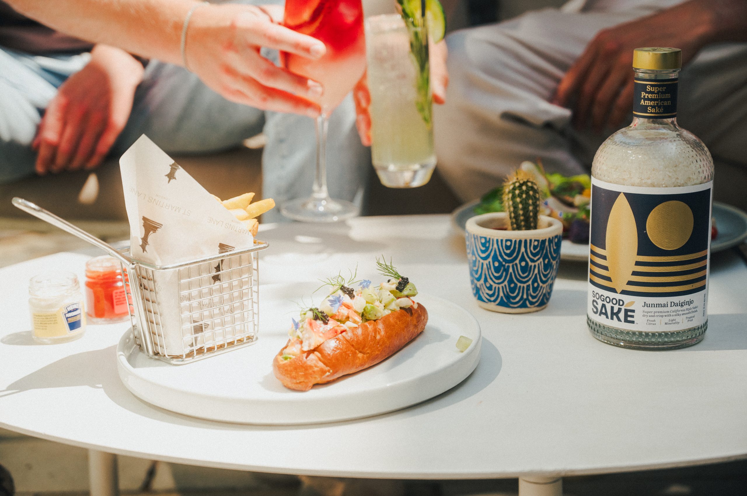Image of a table and two people sat on a sofa in the background holding two cocktails. On the table there is a white plate with a lobster roll and fries, a bottle of SOGOOD SAKE and a small decorative cactus.