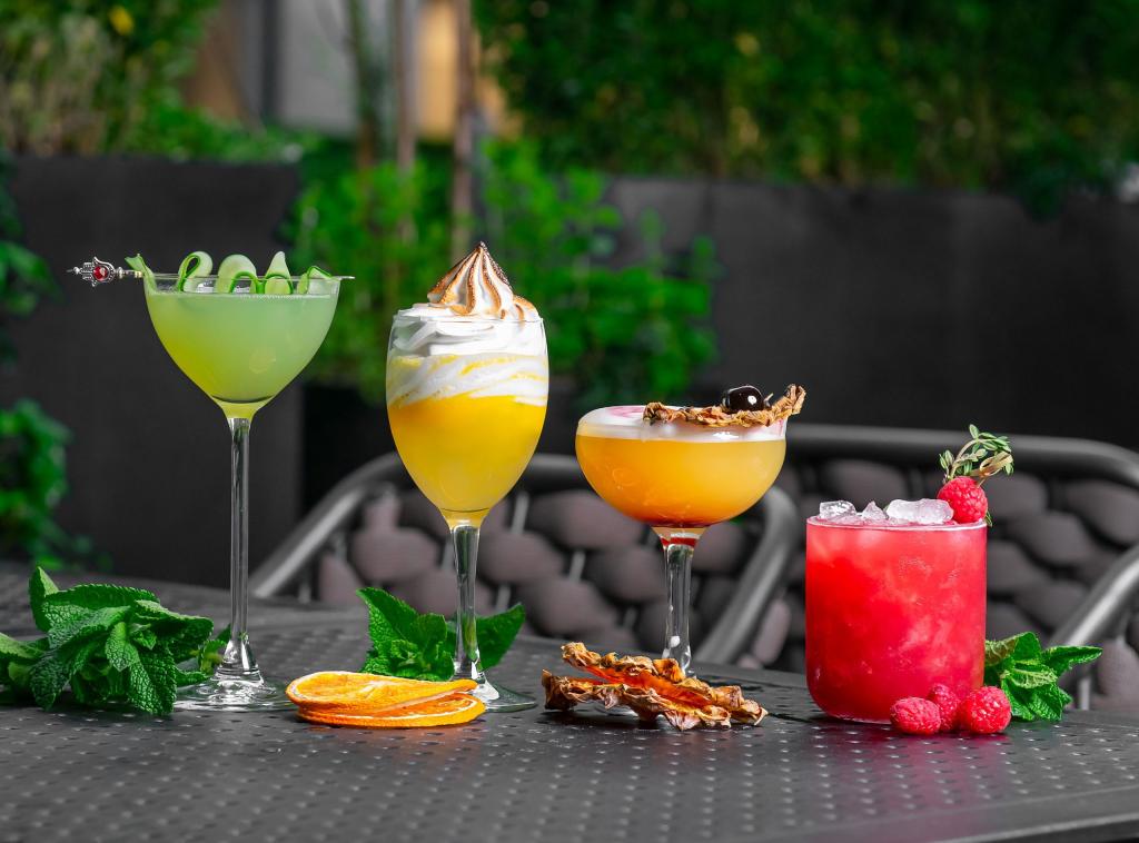 Green cocktail, two orange cocktails, and a red cocktail sit on a dark table on an outdoor terrace with greenery in the background