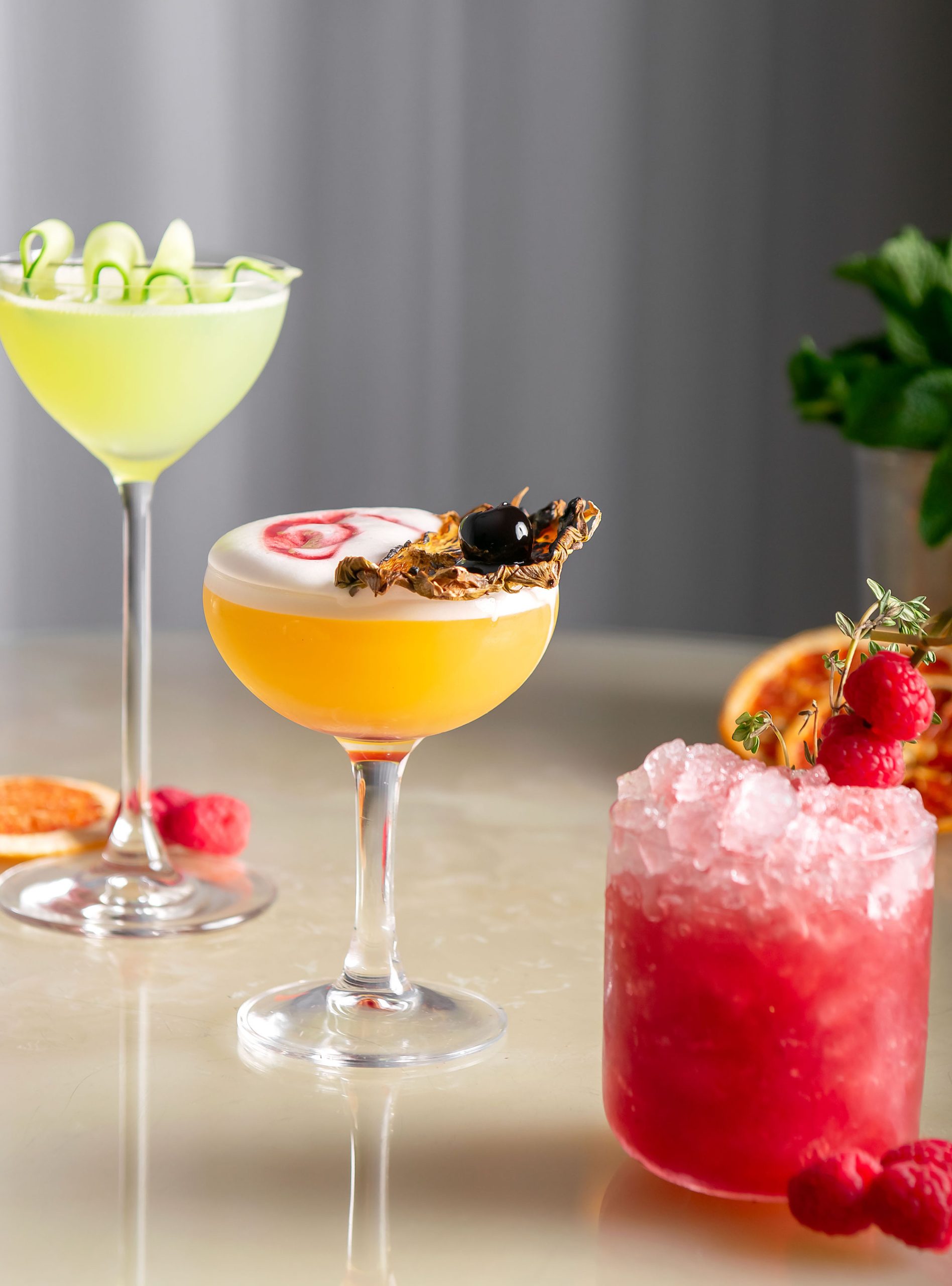 One tall green cocktail, a short orange cocktail, and icey red cocktail all with garnishes sit on beige table