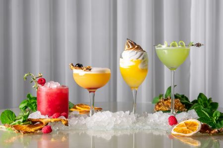 Four elaborate cocktails in red, orange, and green sit on a bar with ice and fruit slices