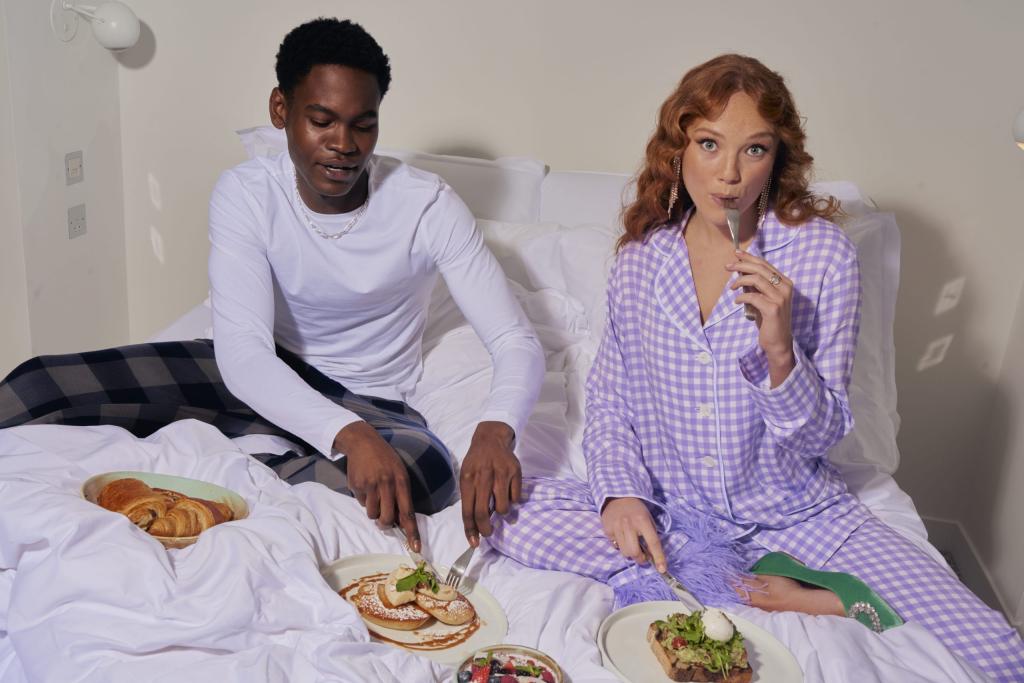 Two people in pyjamas on a hotel bed eating breakfast of four plates some with croissants, pancakes, fruits and avocado and poached egg on toast.