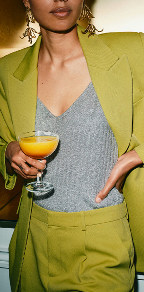 A woman with green suit jacket and trousers holding a bright yellow cocktail. 