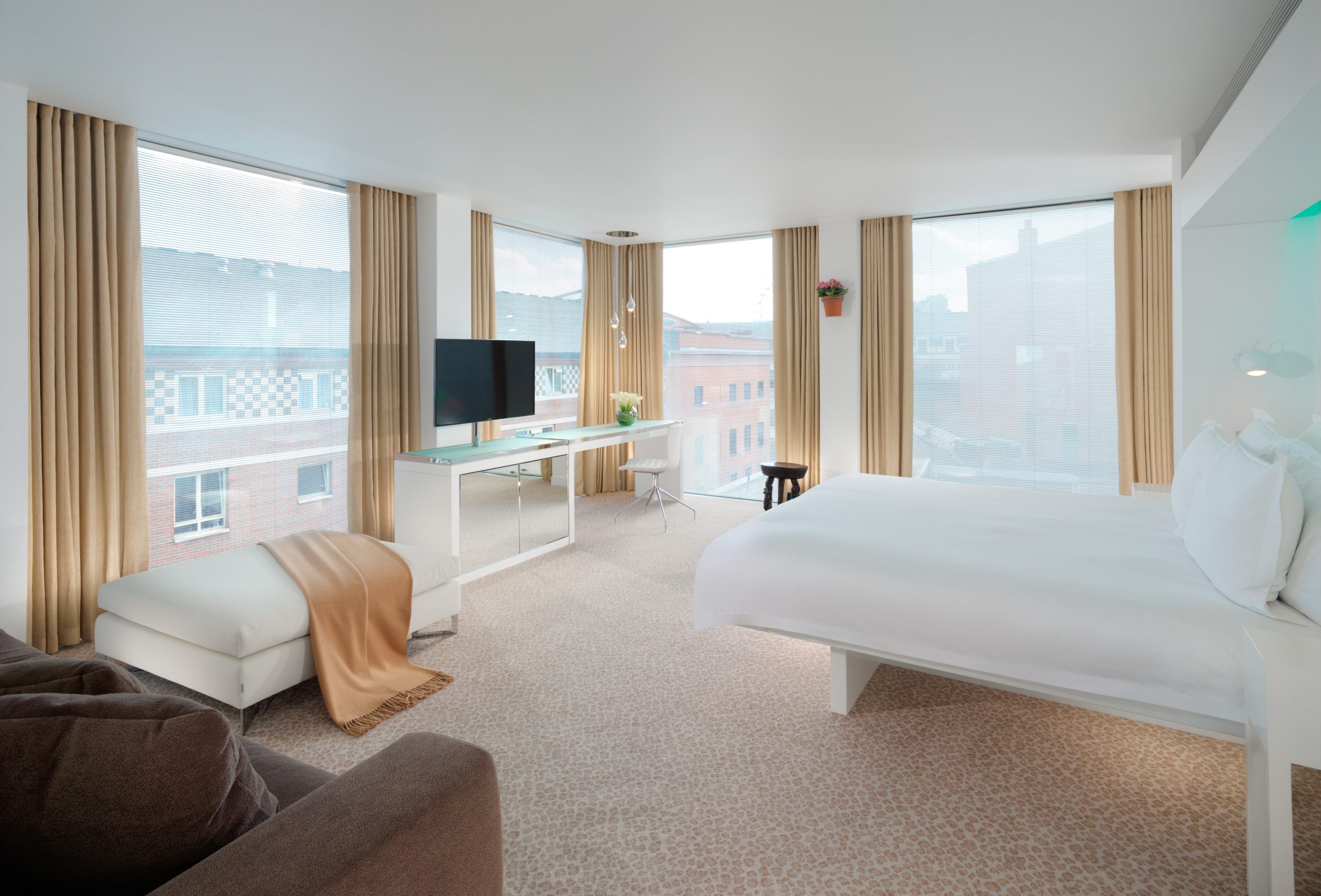 A modern, spacious hotel room featuring large floor-to-ceiling windows with beige curtains, allowing ample natural light. The room is decorated in a minimalist style with a white bed, a white ottoman with a beige throw, and a sleek white desk beneath a wall-mounted TV. The room also includes a small stool, a brown sofa, and a few decorative plants. The carpet has a light, subtle pattern, adding to the clean and bright ambiance of the space. The windows provide a view of a nearby building.