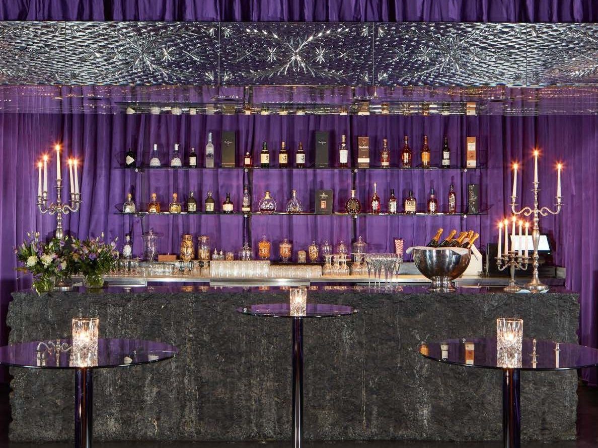 Purple bar backdrop with shelves of liquor bottles, candle sticks, and tall skinny round bar tables with candles